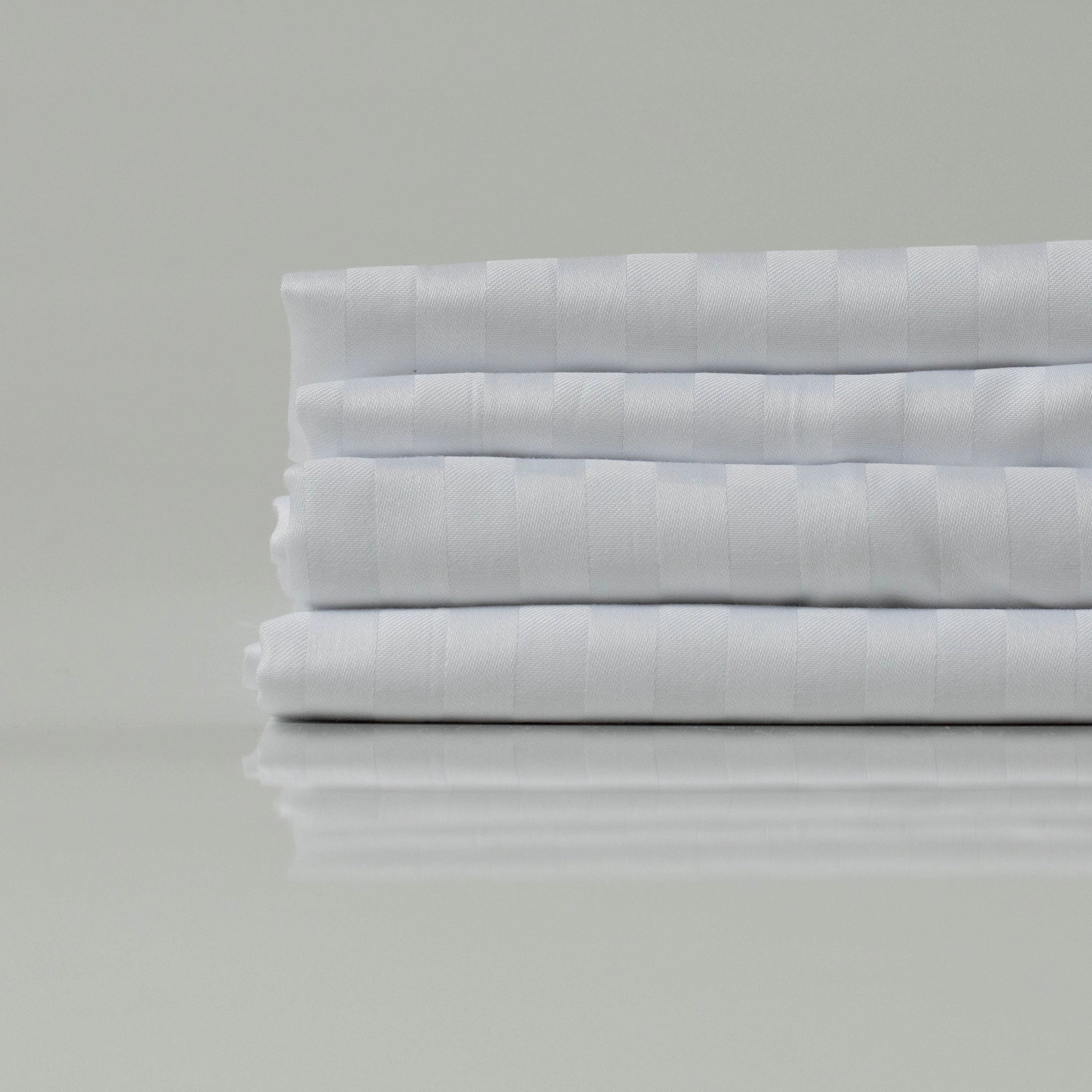 A refined stack of lustrous white bedsheets, folded with precision to reveal their understated stripe pattern and signature sateen sheen—tailored for elevated rest