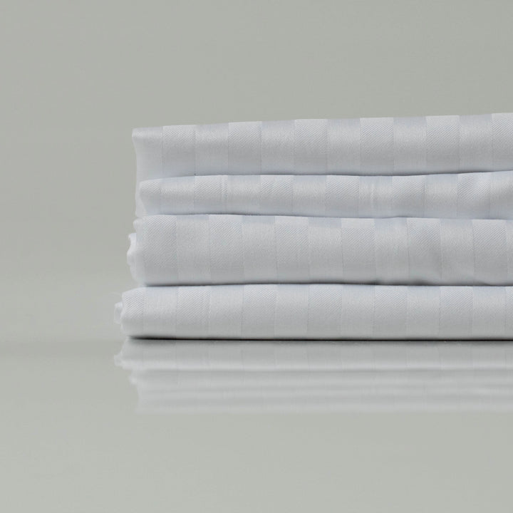 A refined stack of lustrous white bedsheets, folded with precision to reveal their understated stripe pattern and signature sateen sheen—tailored for elevated rest