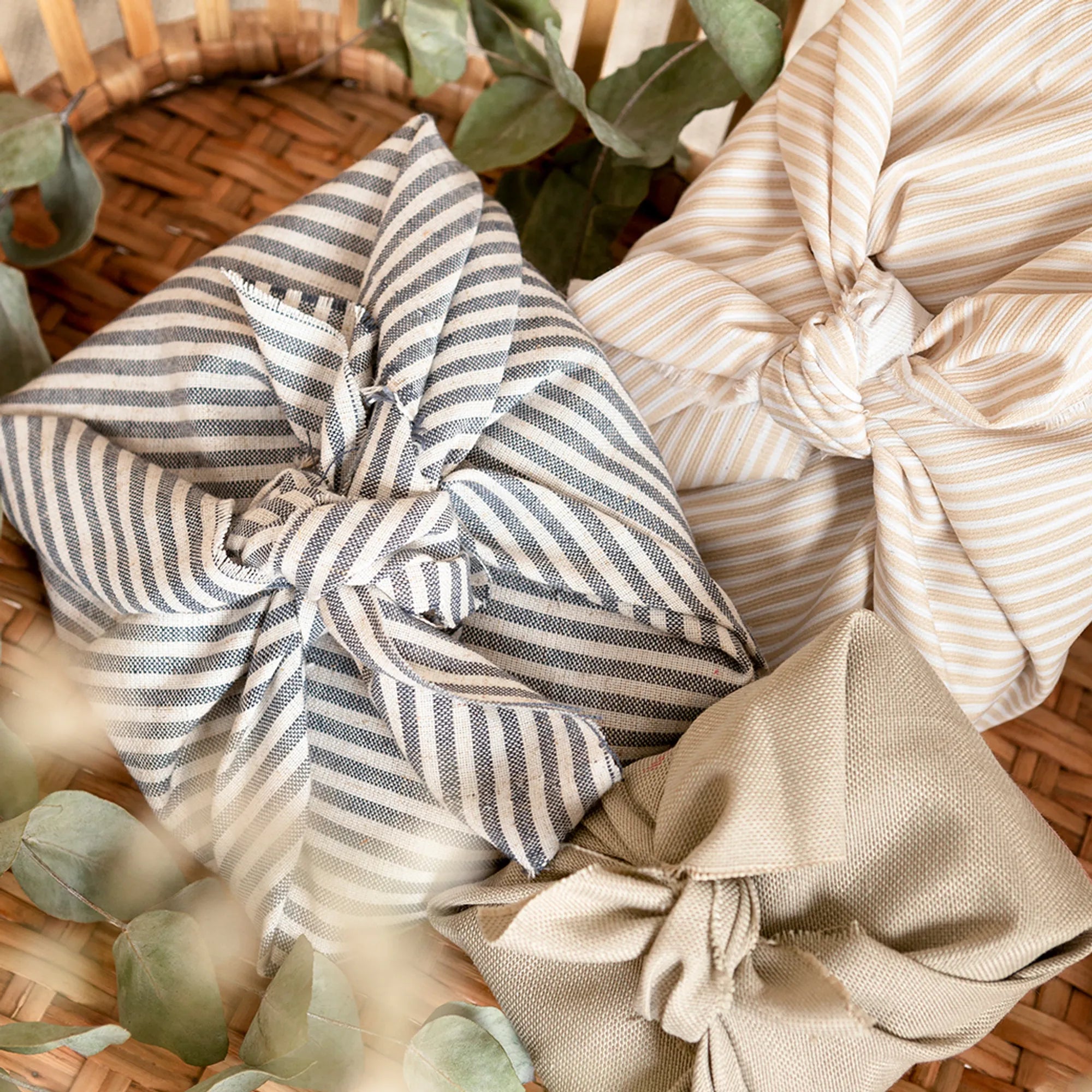 Three beautifully wrapped fabric bundles in earthy stripes and solids, styled in a woven basket for a warm, eco-friendly presentation with a handcrafted touch