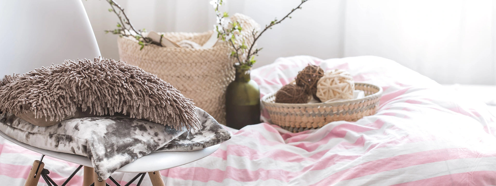 Cozy bedroom scene with a pink and white striped duvet, textured cushions on a chair, and decorative accents in a wicker basket and glass vase