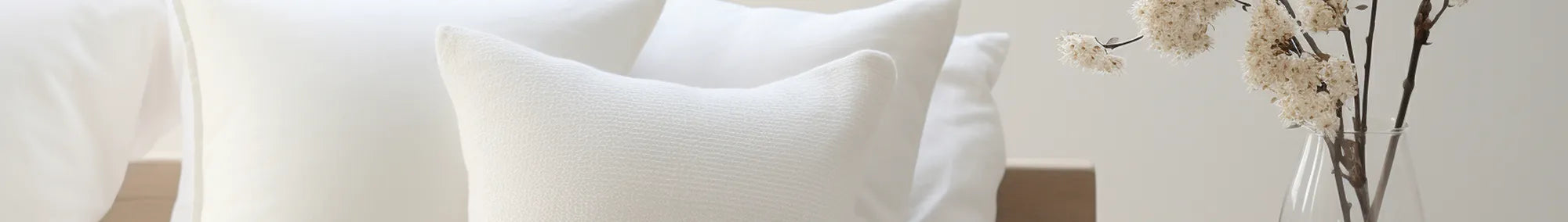 A serene bedroom scene featuring textured ivory pillows and soft white bedding beside a delicate vase of dried blossoms, evoking minimalist charm and eco-conscious elegance