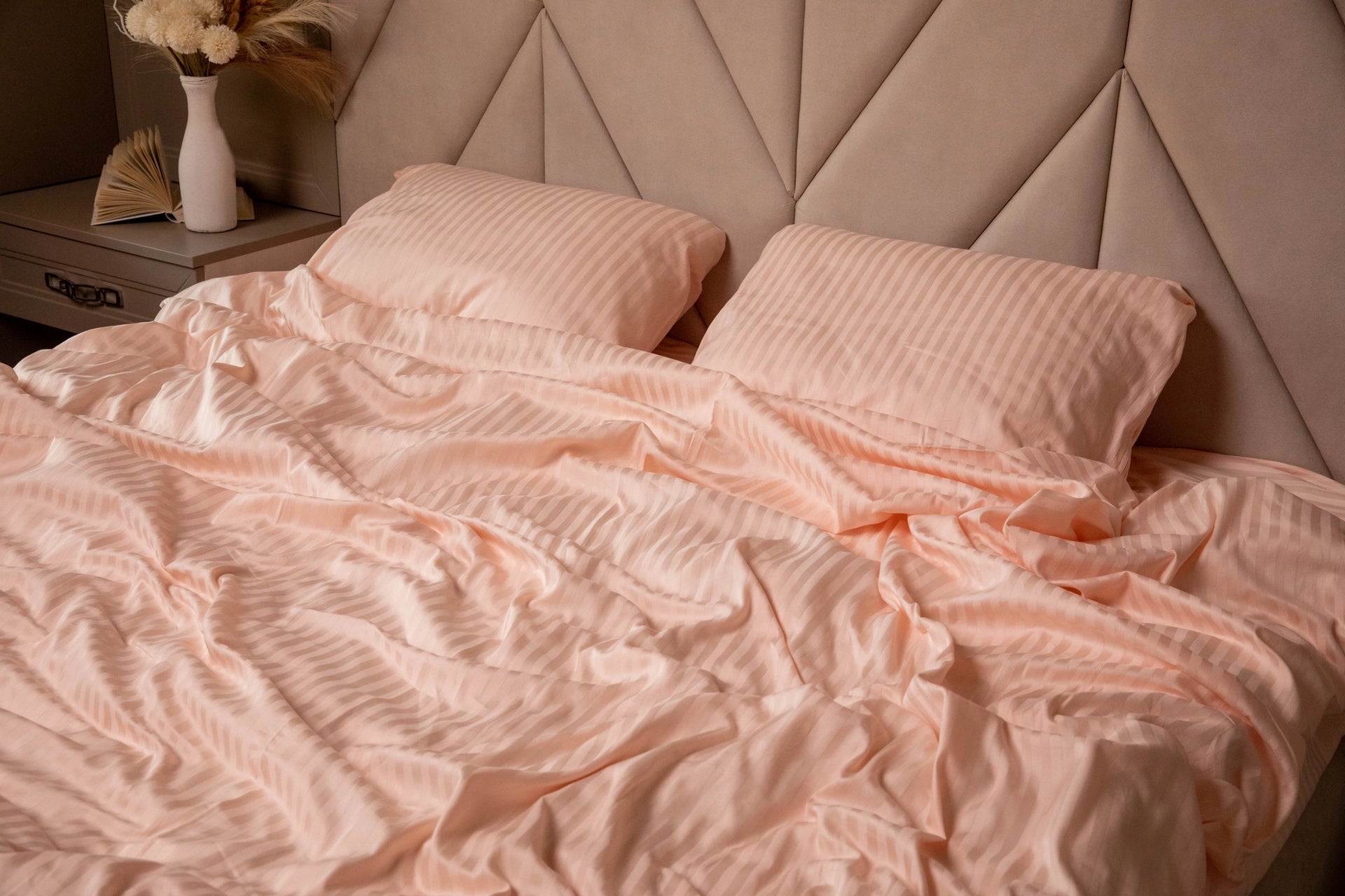 Peach-colored striped bedding on a soft upholstered headboard, styled with a nightstand and neutral floral decor