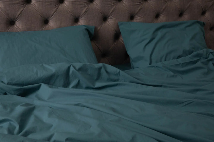 Elegant bed with teal-colored bedding, including a duvet cover and multiple pillows, set against a tufted beige headboard in a classic, softly lit bedroom.